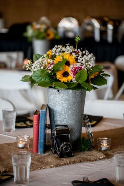 a vintage rustic wedding centerpiece of a bucket with sunflowers and baby's breath, a stack of vintage books and a vintage camera plus candles around
