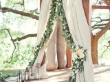 a beautiful ceremony space with neutral curtains decorated with greenery and white blooms and with candle lanterns