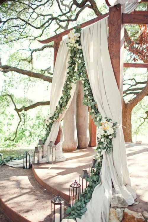 a beautiful ceremony space with neutral curtains decorated with greenery and white blooms and with candle lanterns