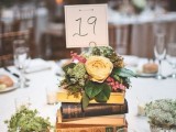 a book stack centerpiece with a bright floral arrangement on top and a table number plus some greenery around