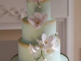 a refined mint green wedding cake with gold edges and luxurious white sugar blooms on a chic stand