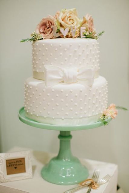 a white polka dot wedding cake with a sugar bow, some pastel blooms on top and greenery is a pretty solution for a spring or summer wedding
