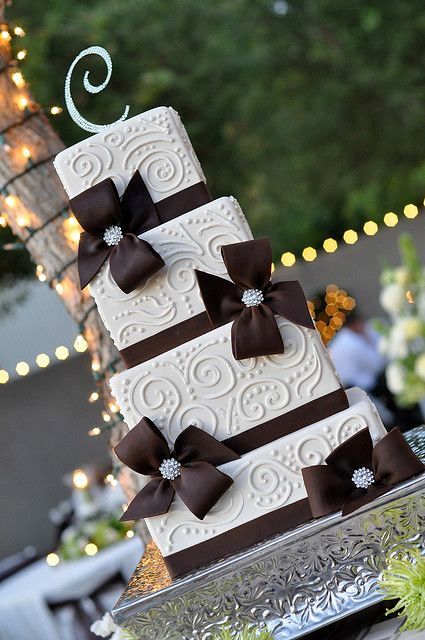 a white patterned square wedding cake with brown ribbons and bows, with embellished brooches and a monogram topper for a chic and refined wedding