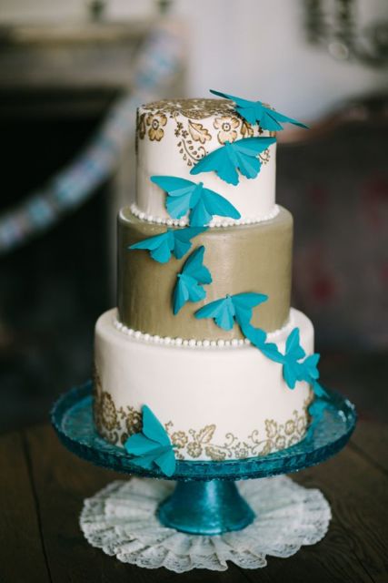 a white and gold wedding cake decorated with blue butterflies and gold floral patterns painted on the piece is a bright and chic idea