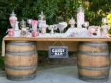 a rustic candy bar made of barrels, a tabletop, a chalkboard sign and lots of pink and white candies in jars