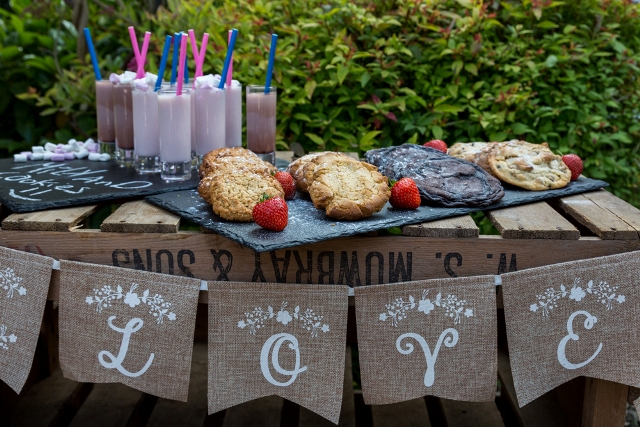 a small cookie bar ona  wooden crate, with a burlap banner, elegant stone trays with cookies and berries and milkshakes