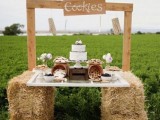 a rustic cookie bar with a tabletop placed on hay, wooden planks and a sign over it, jars with cookies and a wedding cake