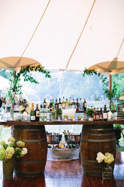 a wedding drink bar with wine barrels and a countertop, greenery, white hydrangeas and umbrellas over it is a lovely idea