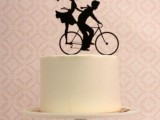 a white buttercream wedding cake with a black silhouette cake topped of a couple riding a bike is a fun and cool idea for a modern wedding