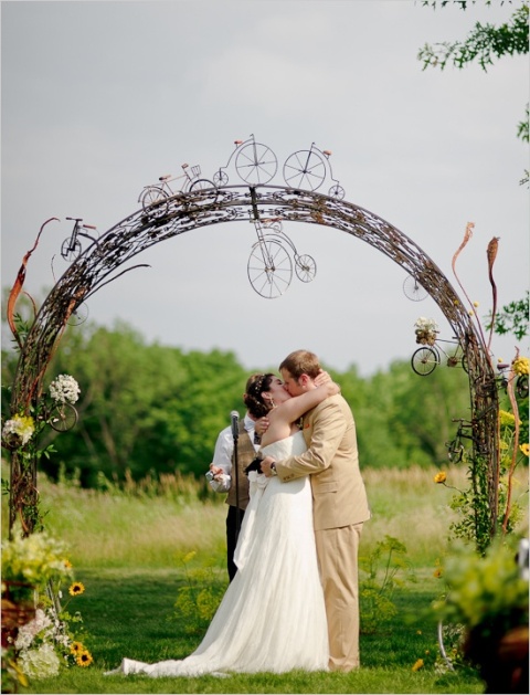 a creative vine wedding arch decorated with white and yellow blooms, greenery and mini bikes to show that the couple loves cycling