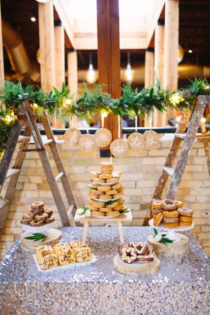 a sequin covered table with donuts on trays and plates, cookies and cupcakes and a greenery and light garland over the sweets table