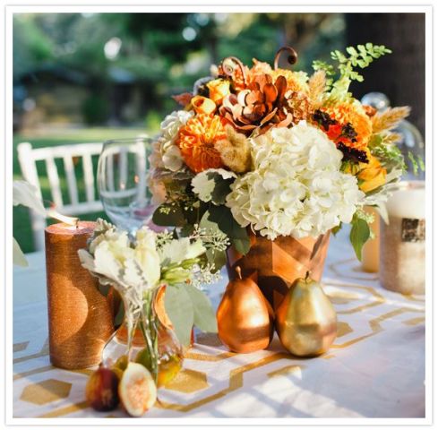 a fall wedding centerpiece of a rust vase, rust and white blooms, greenery and gilded pears, candles around is a truly fall wedding arrangement