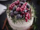a white buttercream wedding cake topped with greenery and frozen berries and cinnamon sticks