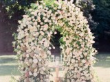 a lush wedding arch covered with greenery and blush blooms is a stylish and romantic idea for a pastel wedding