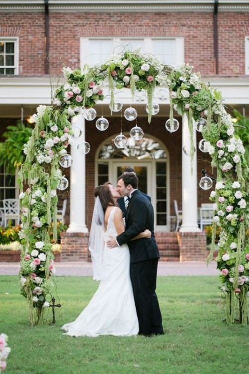 a catchy wedding arch covered with greenery, white and pink blooms and candles hanging in bulbs is wow