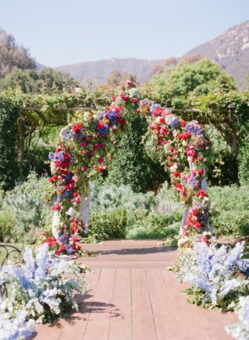 a colorful floral wedding arch with lilac, pink, red blooms and greenery and blue floral arrangements lining up the aisle