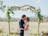 a delicate rustic wedding arch with neutral fabric, greeneyr and white blooms and tree stumps with bottles