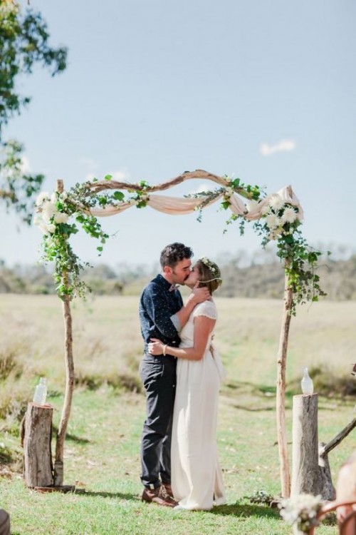 a delicate rustic wedding arch with neutral fabric, greeneyr and white blooms and tree stumps with bottles