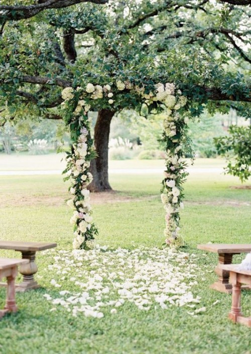 a wedding arch covered with greenery and white blooms and white petals on the ground is a pretty rustic wedding decor idea