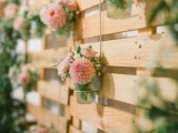 a pallet backdrop with sheer vases and greenery and pink floral arrangements can be used for ceremonies, cake tables, photo booths and so on
