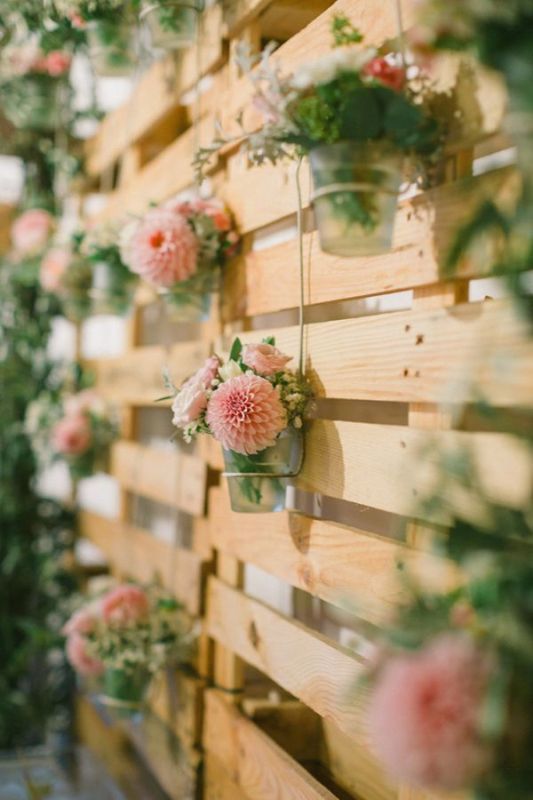 A pallet backdrop with sheer vases and greenery and pink floral arrangements can be used for ceremonies, cake tables, photo booths and so on