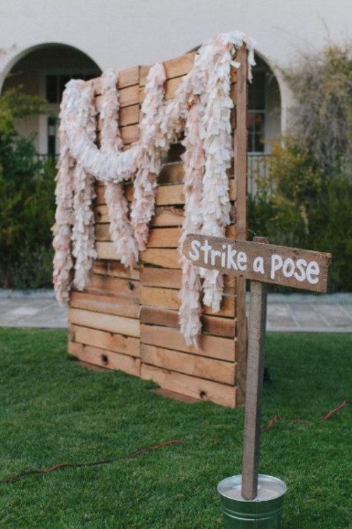 a photo booth backdrop made of pallets with blush and white paper garlands is an easy and eco-friendly idea