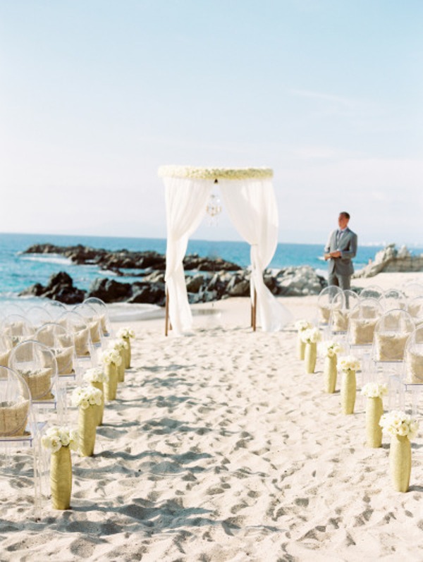 A chic beach wedding ceremony space with acrylic chairs, white blooms in tall vases and a breezy wedding arch decorated with fabric