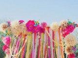 a bright wedding arch covered bold paper pompoms and fans, with long ribbons and polka dot garlands is amazing