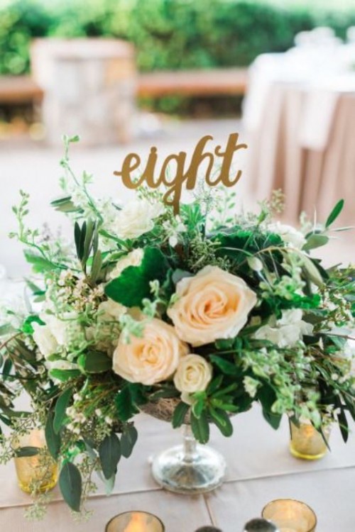 a lush wedding centerpiece with lots of textural greenery and neutral blooms and a calligraphy table number topper