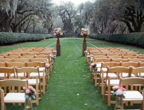 a refined garden wedding ceremony space with a delicate altar with floral arrangements, neutral chairs and petals on the path