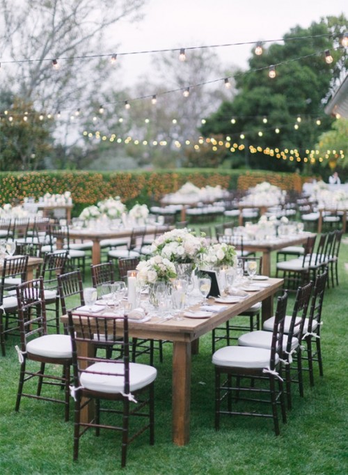 a refined vintage-inspired vintage wedding reception space with wooden tables and neutral chairs, with neutral floral arrangements and string lights over the space