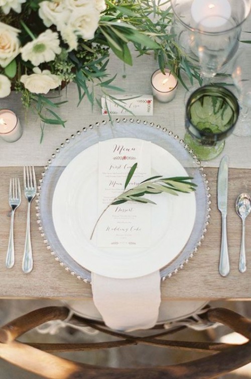 a delicate organic wedding tablescape with a neutral runner, a clear charger, green glasses, a greenery and white flower centerpiece plus candles around