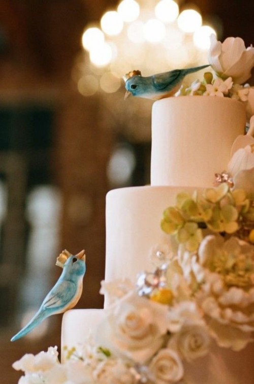 a whimsical wedding cake in white, with sugar blooms, with blue love birds in crowns shows off beauty and romance