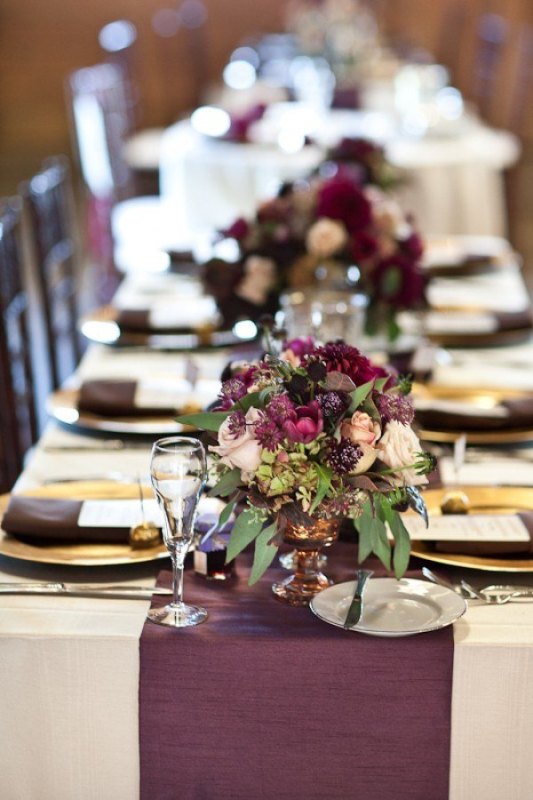 an elegant wedding tablescape with gold chargers, purple and blush blooms and greenery, plum colored napkins and a table runner