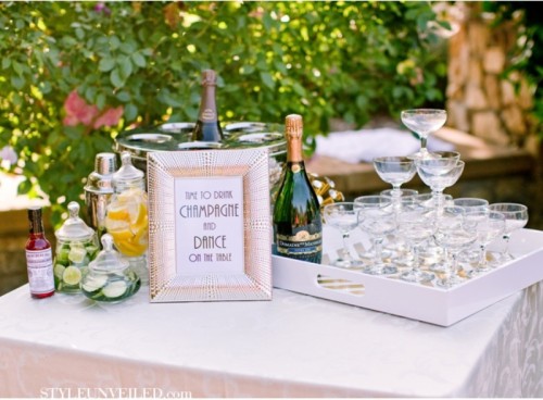 a chic drink table with a mini champagne glass tower, some signs, wine and other alcohol and some fresh fruit slices in jars