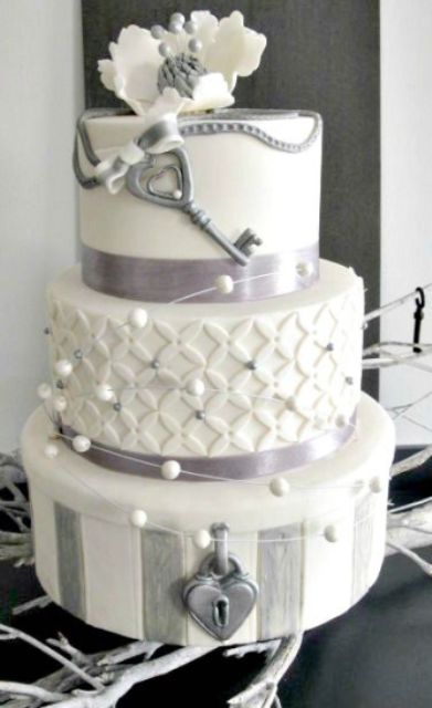 a white and grey wedding cake with plain and patterned tiers, with beads, chain, stripes and a white sugar bloom plus a grey vintage key of sugar