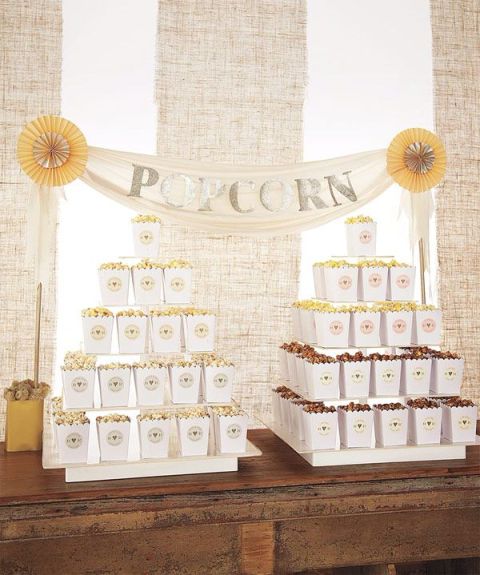 a simple and rustic popcorn bar with a stained table and stands with usual and chocolate popcorn in paper bags and a garland over it
