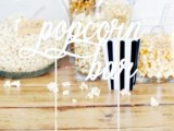 a simple and modern popcorn bar with a sign, glass bowls and jars with popcorn and striped paper bags
