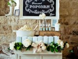 a simple rustic popcorn bar with  a vintage cabinet dressed up with a lush greenery and bloom garland with a bow, a bucket with popcorn, condiments and paper bags