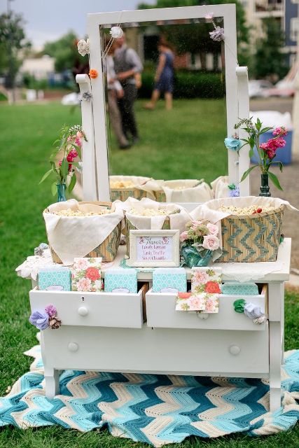 a simple popcorn bar with a vintage dresser that features woven baskets with popcorn and paper bags to store it plus some blooms