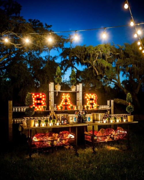 a gorgeous wedding drink bar composed of open shelving units and tables, with marquee letters, greenery decor and candles is a fantastic solution and it looks amazing at night