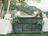 a vintage-inspired wedding bar of a grey sideboard, a couple of tables, greenery and lots of drinks and glasses on all the parts