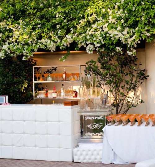 a lovely outdoor drink bar with a bar stand covered with white upholstery, some open shelves with bottles and glasses, a wide cooler and a table with drinks