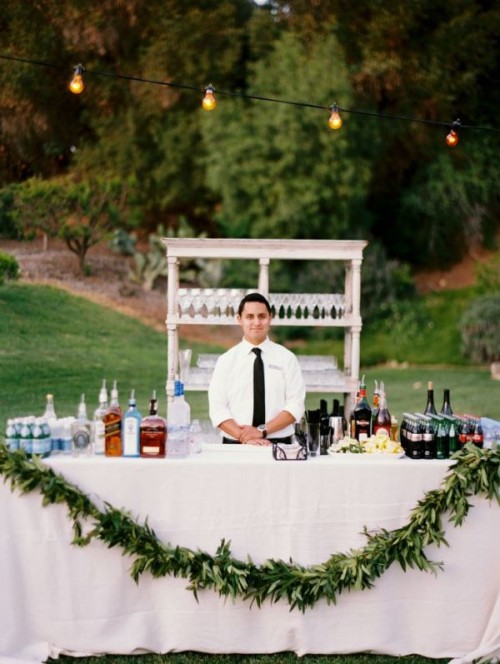 a lovely wedding drink bar - a table with white tablecloth and a greenery garland, an open shelving unit with glasses is a lovely idea to go for