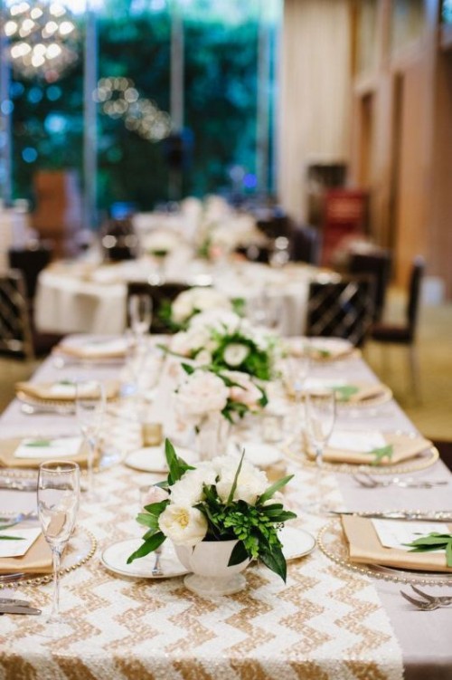 a lovely neutral wedding tablescape with a chevron sequin table runner, white blooms and greenery, clear plates and gold napkins