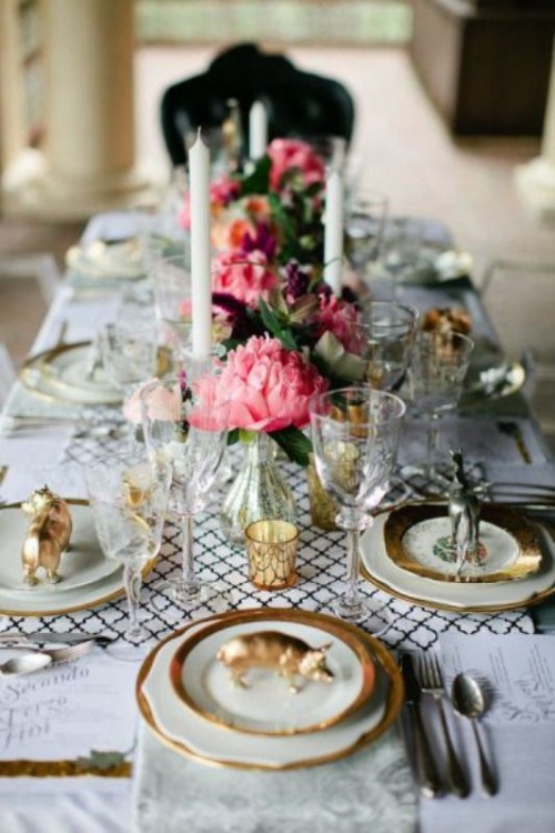 a beautiful wedding table setting with a white tablecloth and printed runners, gold-rimmed plates, candles and pink peonies as centerpieces