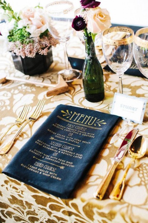 a refined wedding tablescape with a gold and white printed tablecloth, neutral blooms and greenery, a black menu and gold cutlery