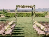 a refined backyard wedding ceremony space with a beautiful green iterwoven arch with blooms and refined chairs with blooms and greenery