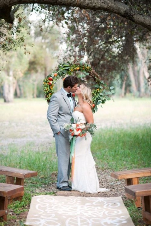 a pretty rustic backyard ceremony space with a lovely rustic wreath hanging from a branch, with wooden benches in rows is very cute