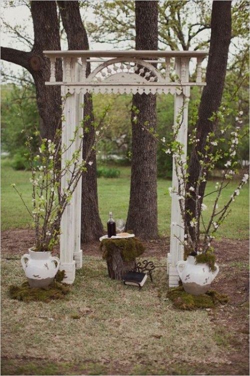 a vintage backyard wedding arch of carved wood painted white, with blooming branches in jugs and moss on the ground for a spring backyard wedding